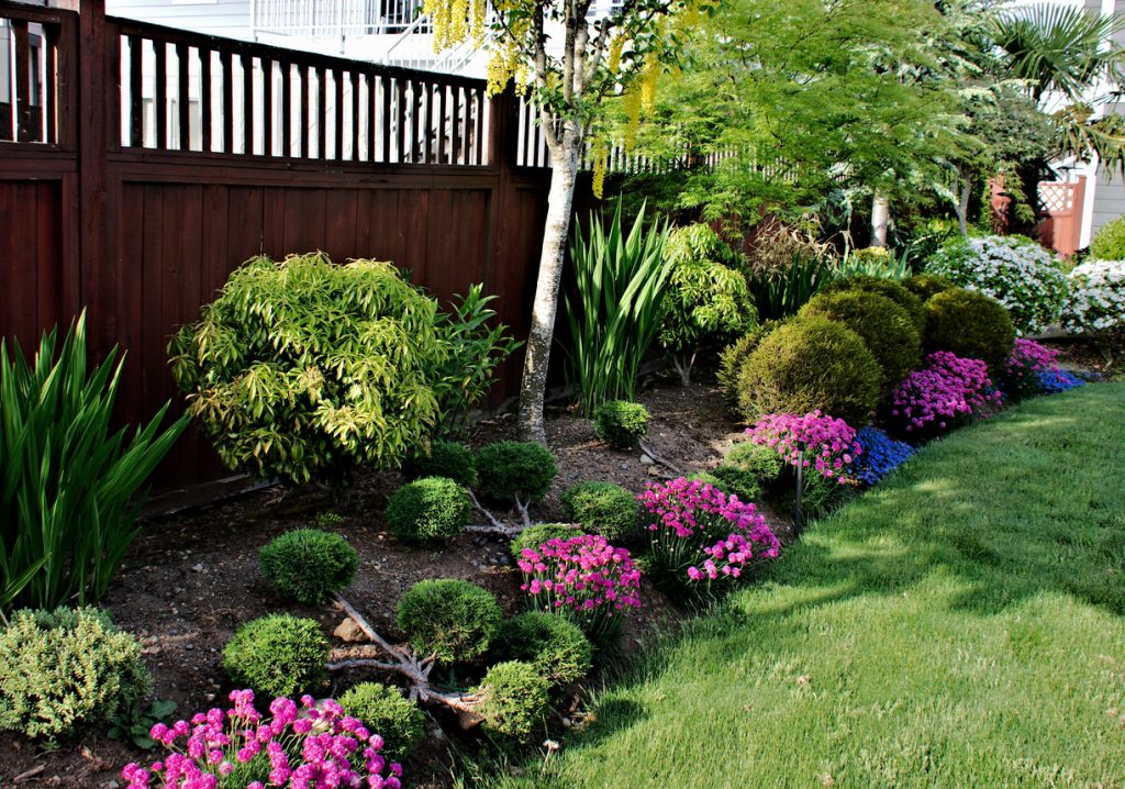 A flowerbed sandwiched between a green grass area of lawn and a brown colored wooden fence consisting of a blend of bushes and flowers in bloom as well as trees in a formal setting.